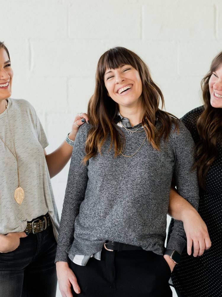 3 women smiling and standing beside white wall
