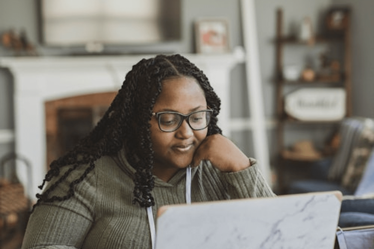 A woman with braids and glasses sitting at a table, working on a laptop.
