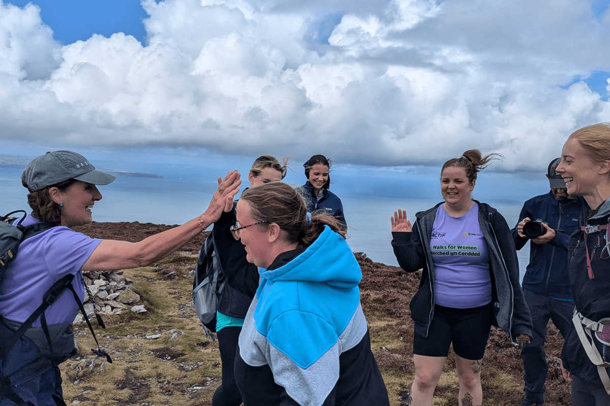 A group of six people stand on a rocky hillside overlooking the coast, with one person raising a hand for a high-five. Heather-covered moorland and dramatic clouds fill the background.
