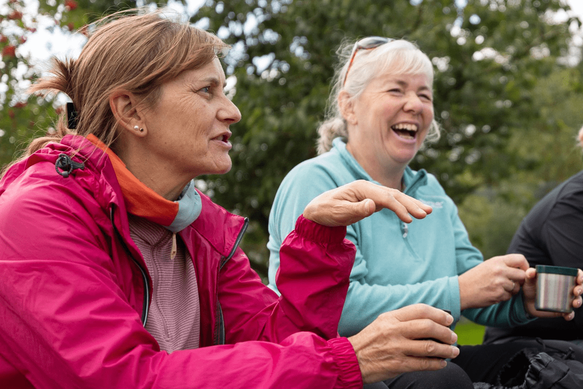 Two people in outdoor clothing sit together outside, with one person in a bright pink jacket speaking while another holds a mug.