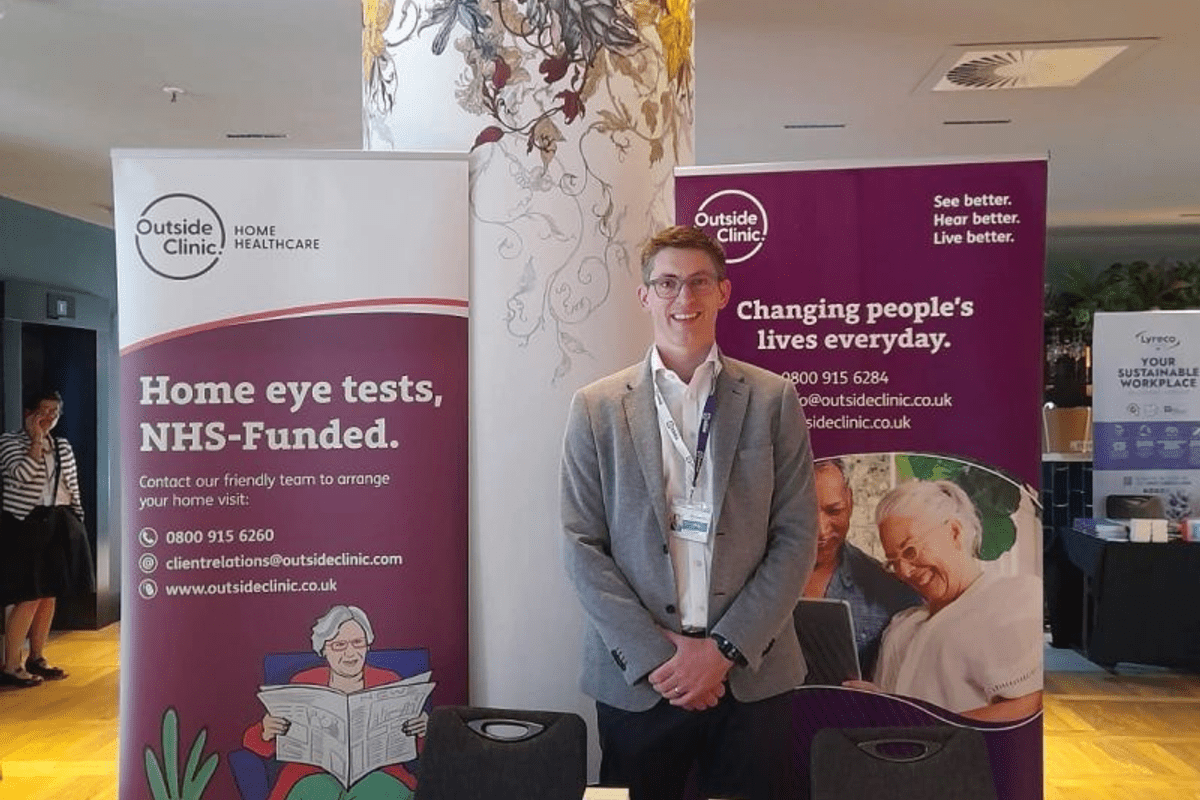 A man, smartly dressed stood in front of OutsideClinic branding and behind a table full of freebies - chocolates, OutsideClinic branded bags, pens.