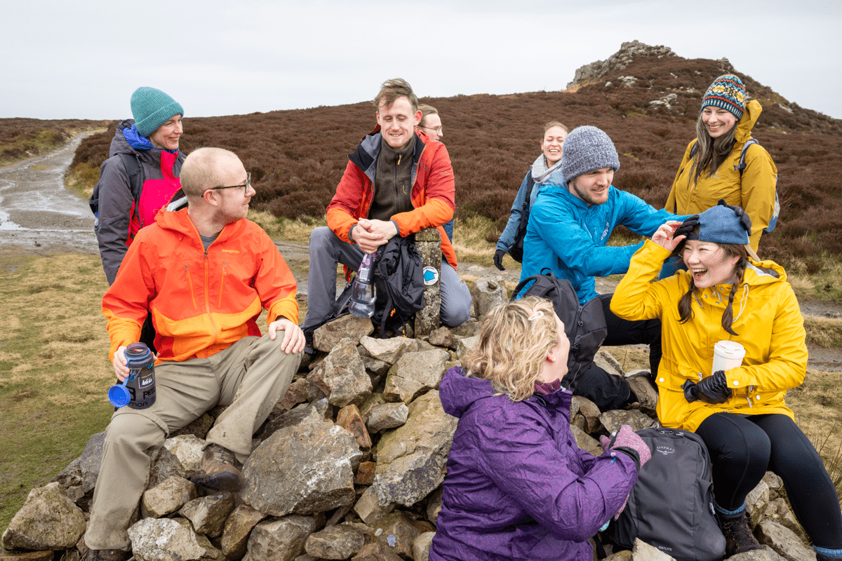 A group of walkers in colourful outdoor clothing sit and stand on a pile of rocks on a moorland hillside, with several people smiling and talking.