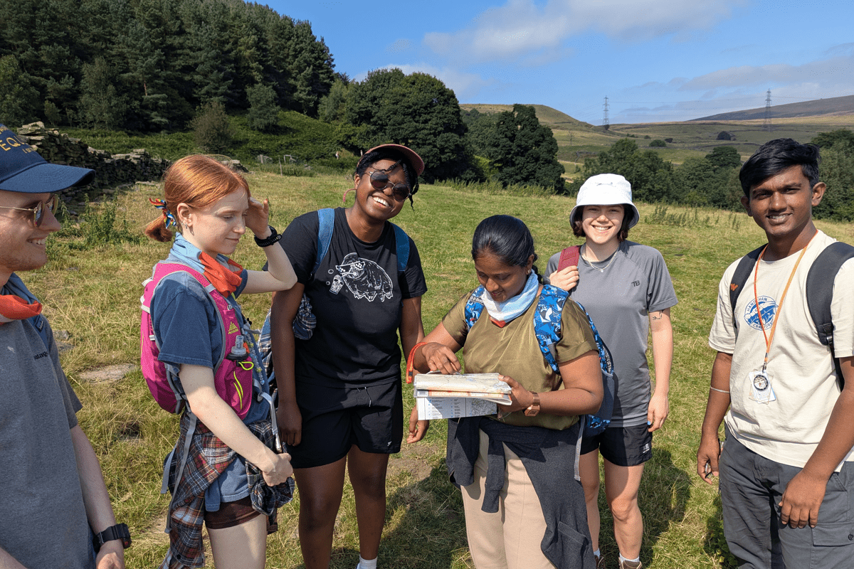 A group of people stand in a countryside landscape, reviewing a map, with hills and woodland in the background.
