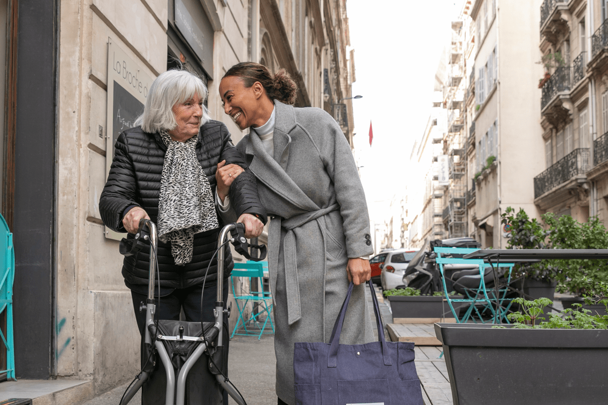 Photo d'une intervenante qui accompagne une senior dans sa promenade en extérieur, elles sourient toutes les deux