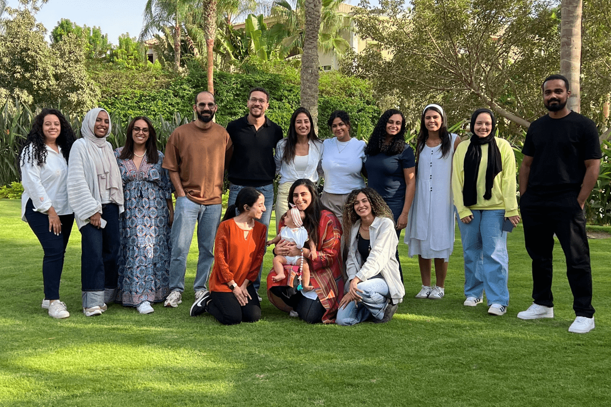 A group of colleagues stand and kneel together on a green lawn under tall palm trees, smiling at the camera with a small child in the center on a bright sunny day.