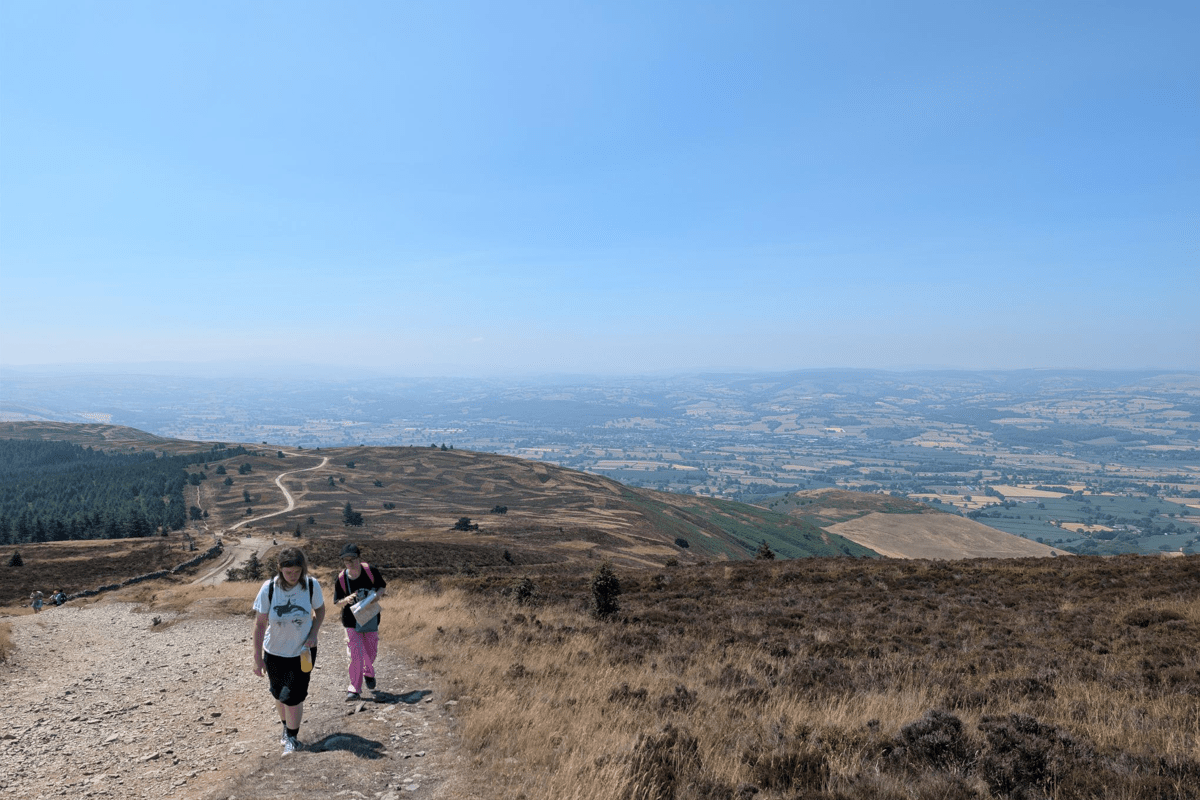 Two people walk along a wide gravel path across open moorland, with rolling hills and patchwork fields stretching into the distance under a clear blue sky.