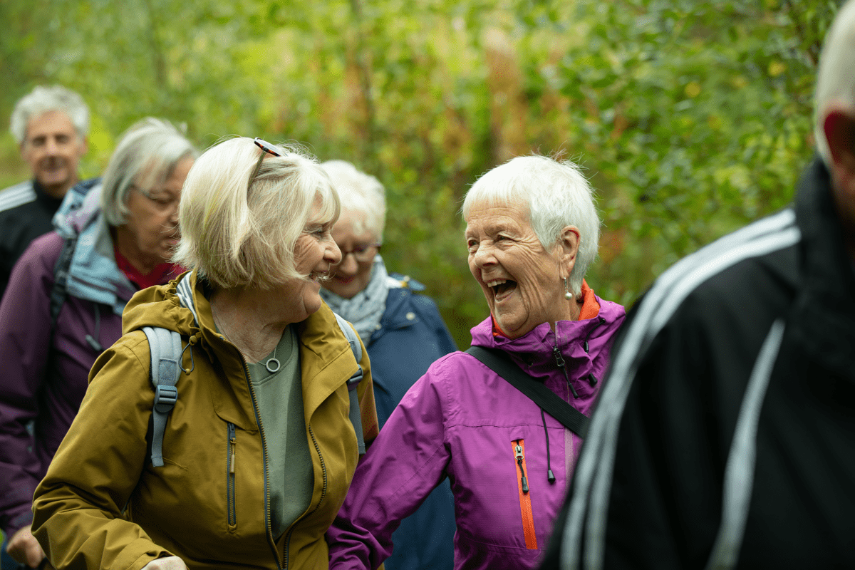 Several people walk together along a wooded path, with two people in the foreground talking as they walk.