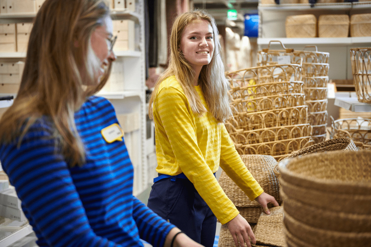 Kaks IKEA töötajat, kellest üks kannab sinise ja teine kollase särgiga vormi, korrastavad poe müügisaalis punutud korvide rida.