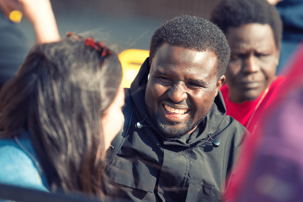 A group of people gathered outdoors in conversation, with one person in a black jacket in the centre facing another person in the foreground.