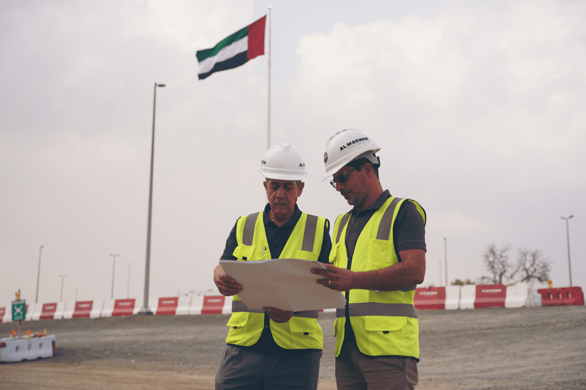 wo construction engineers from Al Marwan Construct wearing high-visibility vests and white hard hats, reviewing a site plan outdoors with the United Arab Emirates flag flying in the background.