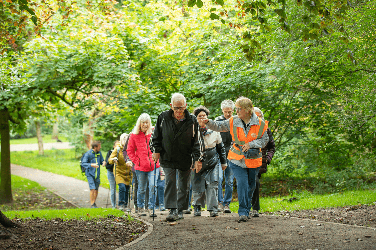 A group of people walk together along a path through a green park, led by a person in a high-visibility vest walking alongside an older person using a walking stick. Trees and dense foliage line the path, with additional walkers following behind.