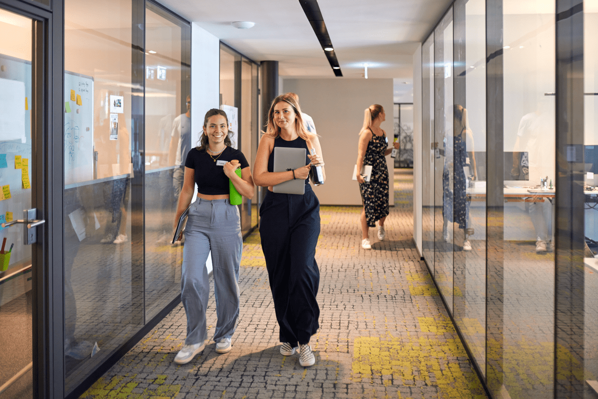 Two women walking and chatting in a modern office hallway with glass-walled meeting rooms