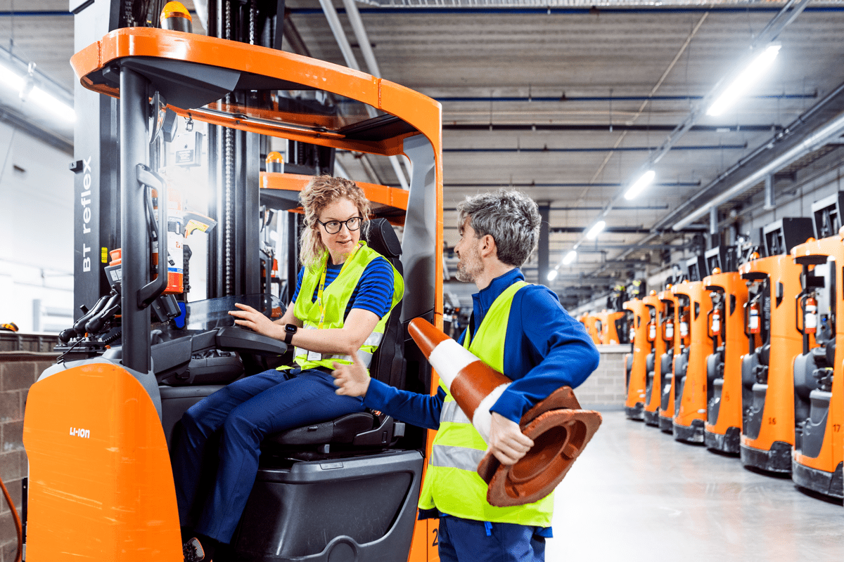 Du IKEA darbuotojai su mėlynos spalvos uniformomis ir apsauginėmis liemenėmis parduotuvės sandėlyje: moteris sėdi už keltuvo vairo, o vyras stovi šalia ir laiko rankoje eismo kūgius.