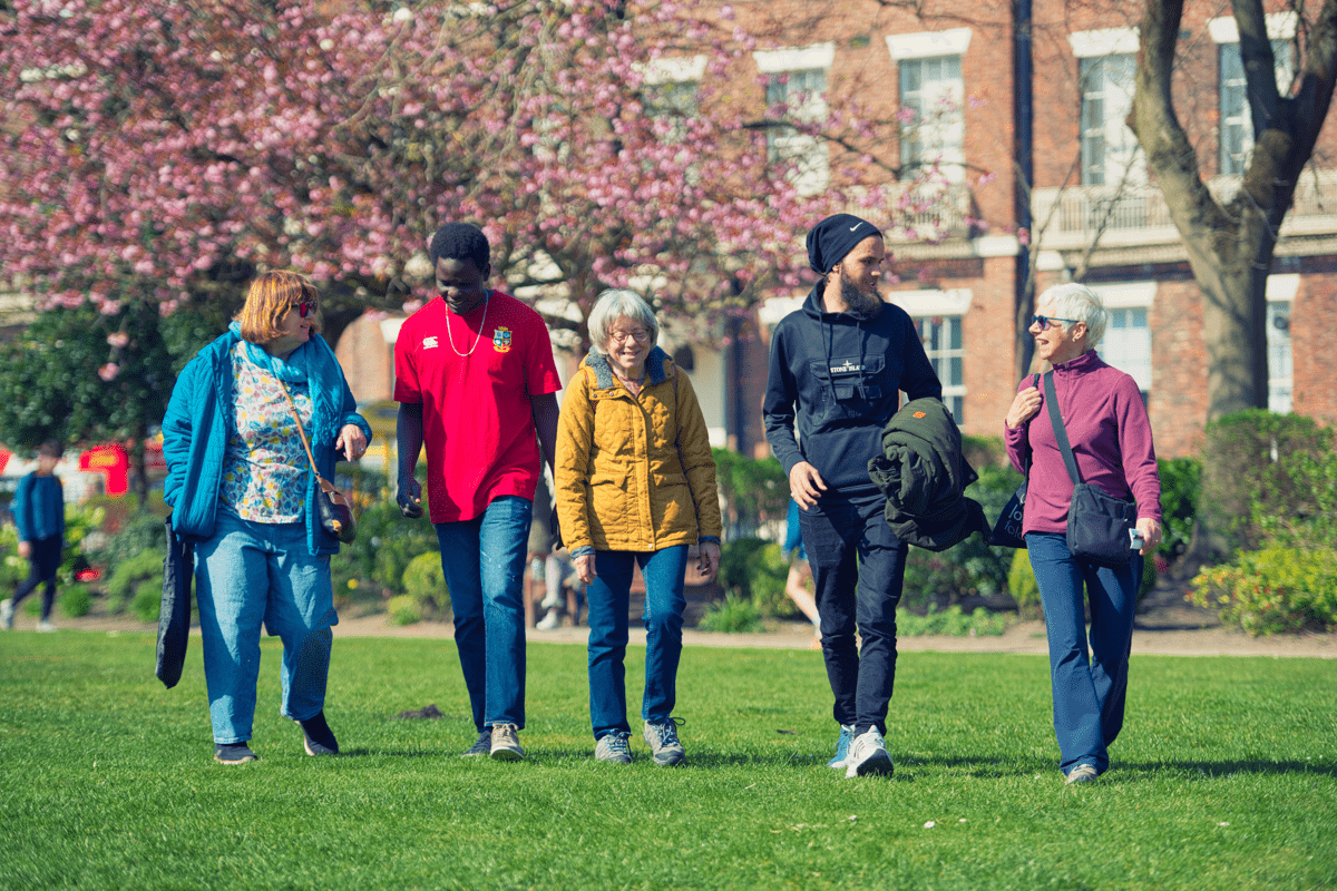 Five people walk together across a grassy park, with blossoming trees and residential buildings in the background.
