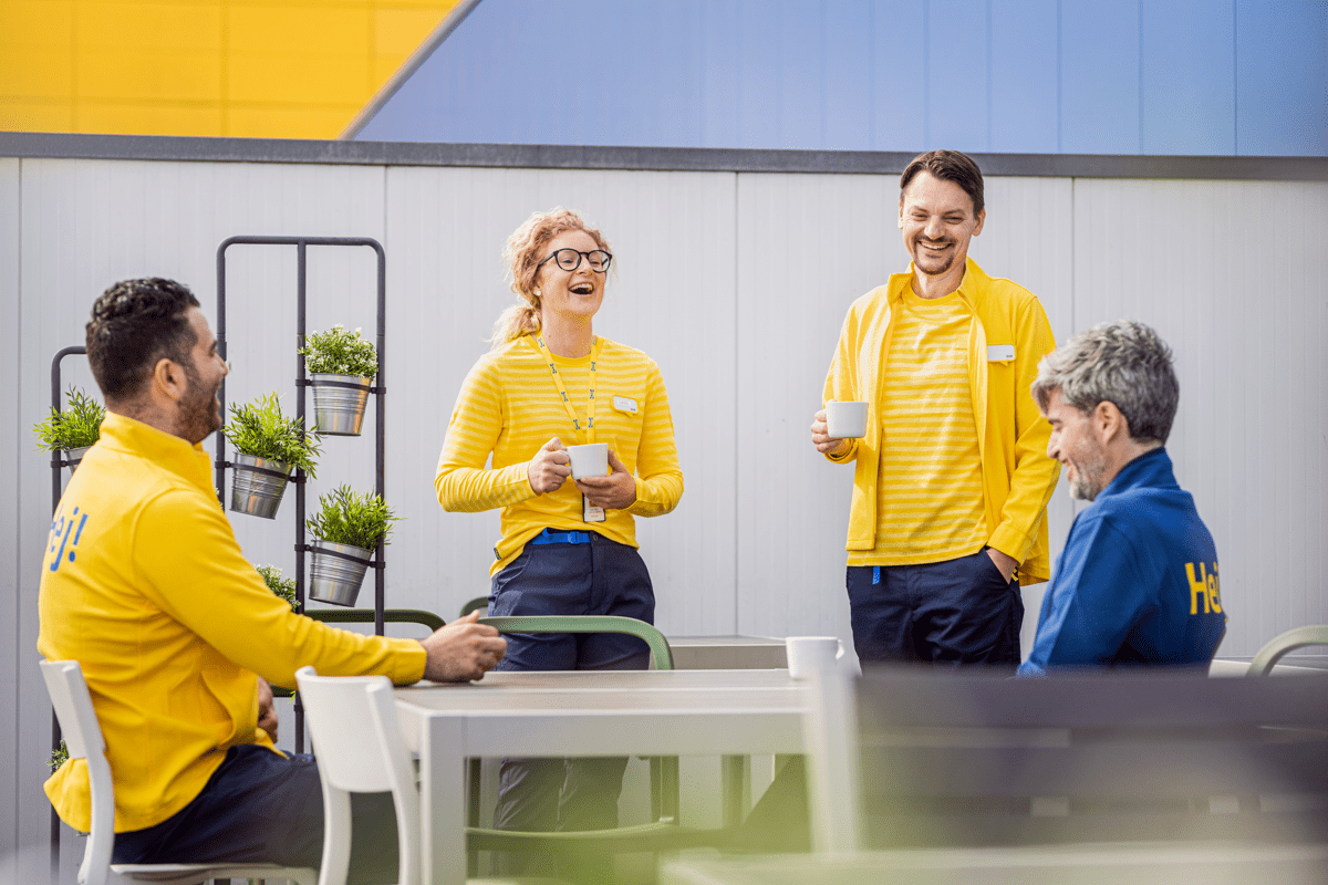 IKEA darbuotojai su uniformomis sėdi ir stovi aplink stalą, šypsodamiesi kalbasi ir laiko rankose puodelius. Atrodo, lyg vyktų neformalus susitikimas arba kavos pertraukėlė „fika“.