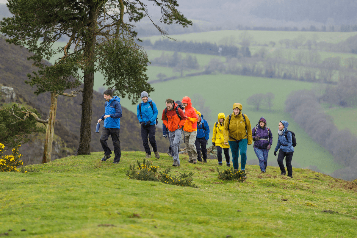 A group of walkers in colourful waterproof jackets hike across a grassy hillside, with a valley and patchwork fields stretching into the distance behind them.