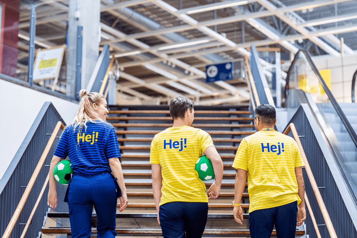 Trys IKEA darbuotojai su geltonos ir mėlynos spalvų marškinėliais lipa parduotuvės laiptais į viršų, o ant jų uniformų nugarų matosi užrašas „Hej!“
