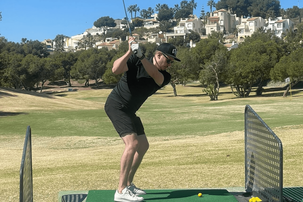 Man spelar golf på grön golfbana med bebyggelse i bakgrunden.