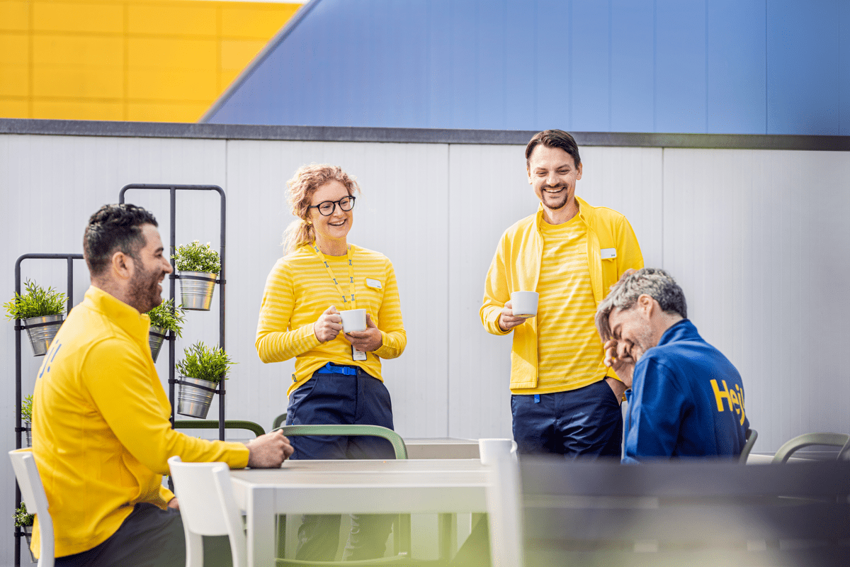 Rühm vormis IKEA töötajaid seisavad ja istuvad ümber laua. Nad naeravad ja hoiavad käes kruuse, millest võib järeldada, et tegu on tiimikoosoleku või fika pausiga. 