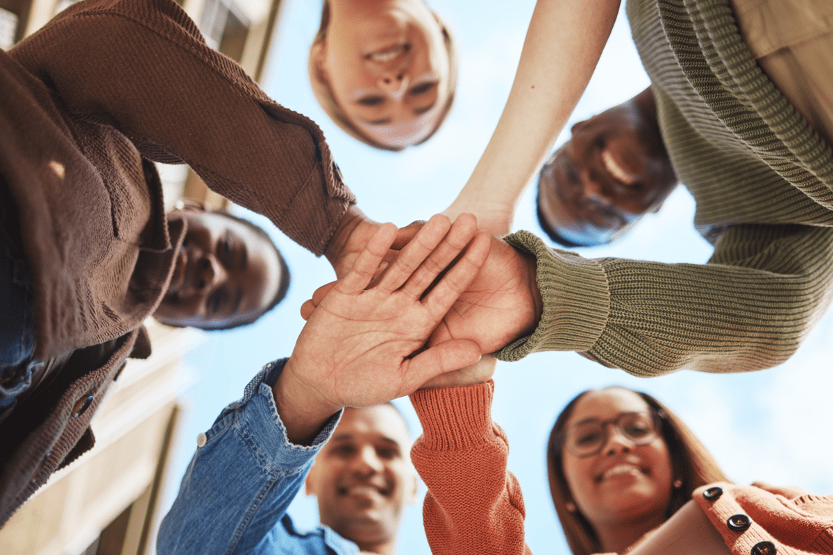 	Fem personer står i en cirkel och lägger sina händer ovanpå varandra, sedda underifrån mot himlen.