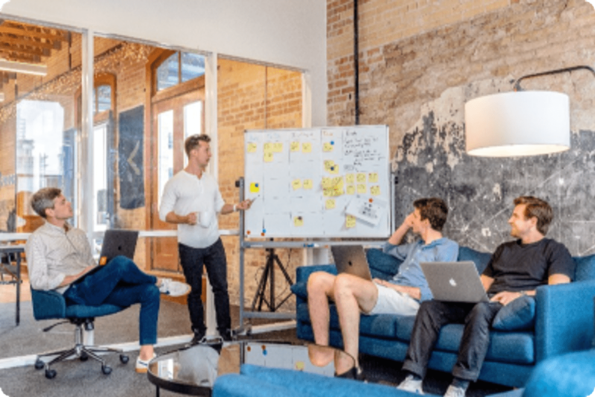 A team of four men collaborating in a modern office with exposed brick walls; one man stands pointing at a whiteboard covered in sticky notes while others watch from a sofa and chair.