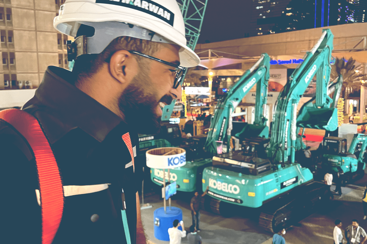 A smiling man in a white Al Marwan branded hard hat looks out from a balcony over a night-time display of turquoise Kobelco excavators and heavy machinery at an outdoor exhibition.