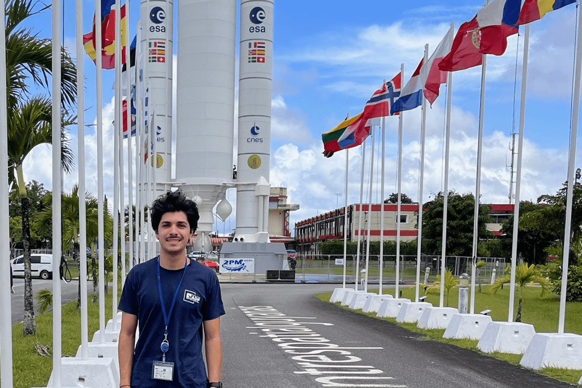Portrait de Calvin ISSART qui pose devant une fusée sur un site spatial, entouré des drapeaux de plusieurs pays.