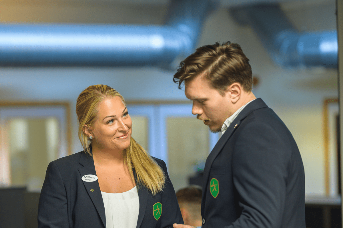 Två kollegor i skoluniform samtalar inomhus, båda bär kavajer med skolans emblem.