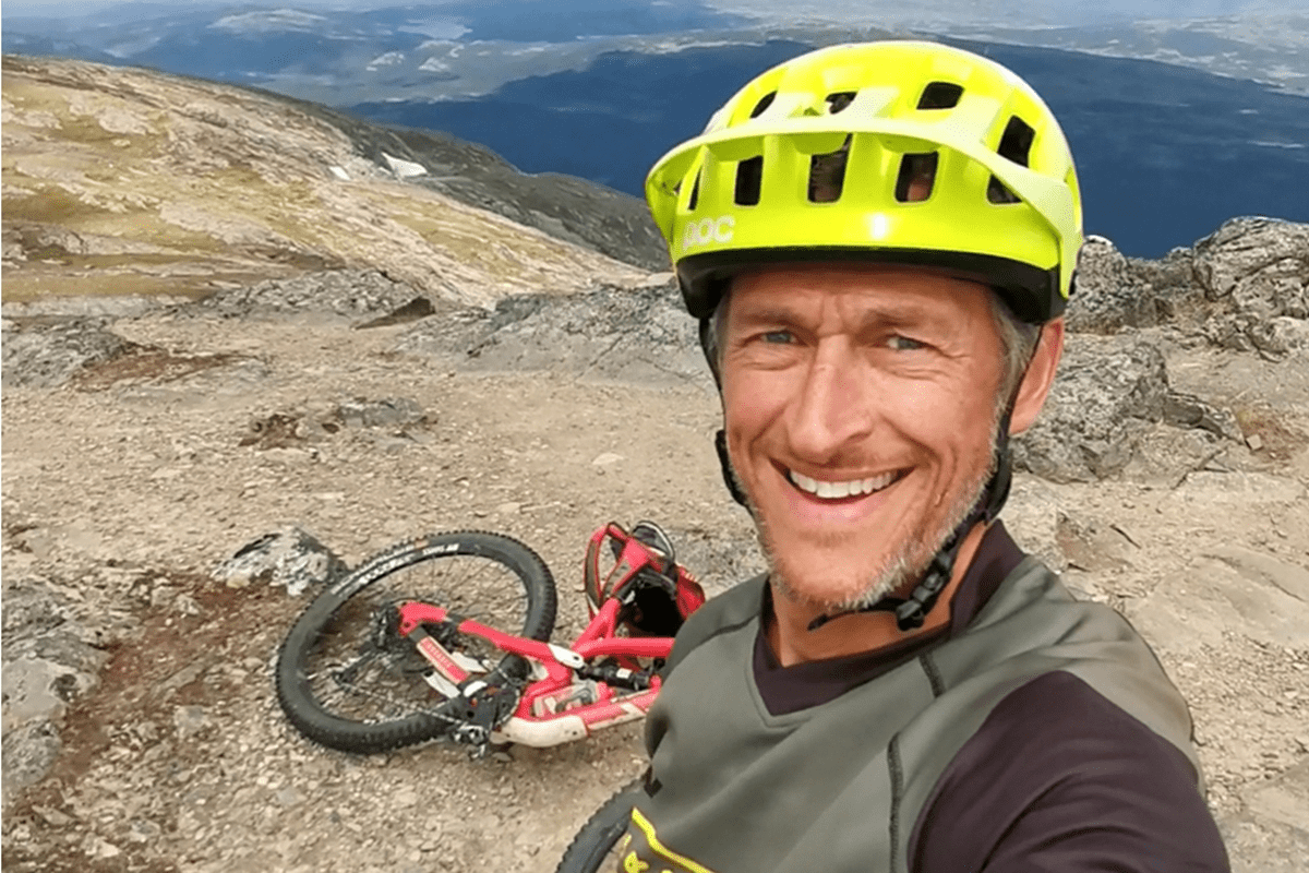 Man med cykelhjälm tar en selfie på ett berg med en mountainbike i bakgrunden.