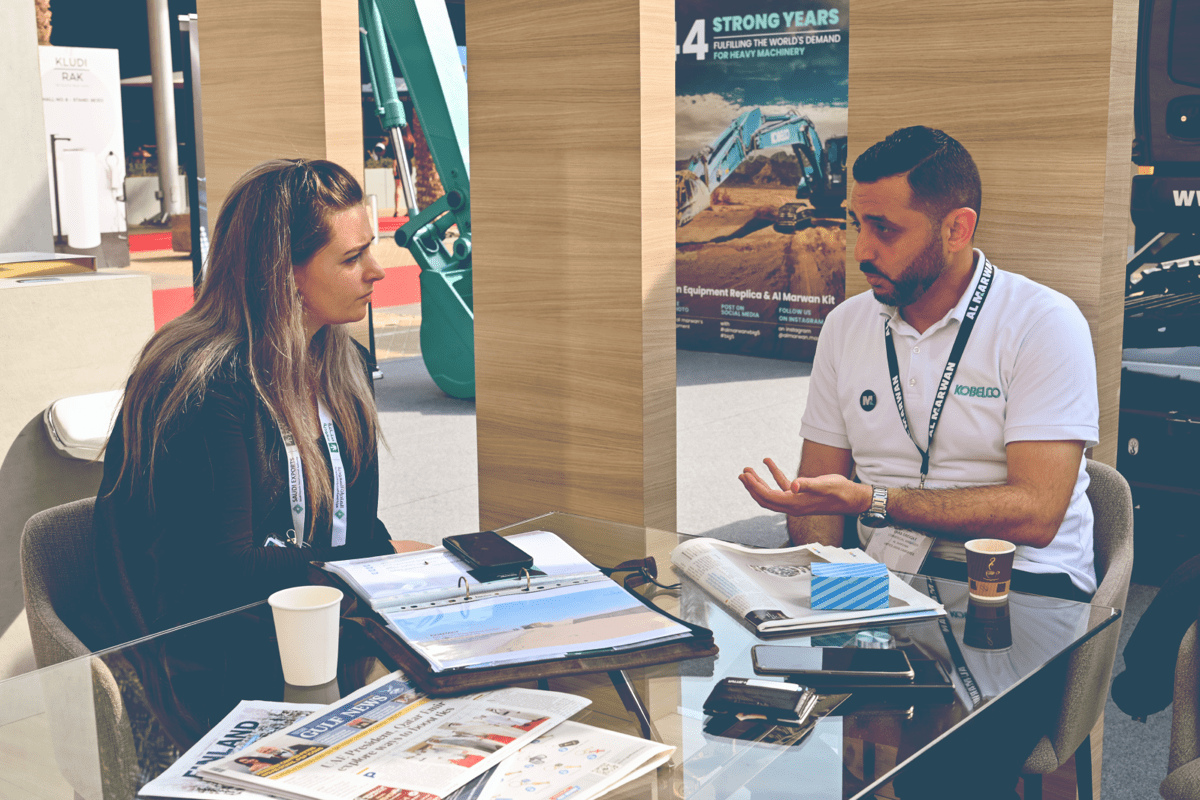 An Al Marwan representative wearing a white Kobelco polo shirt discussing heavy machinery with a female client at a glass table during a construction trade show.