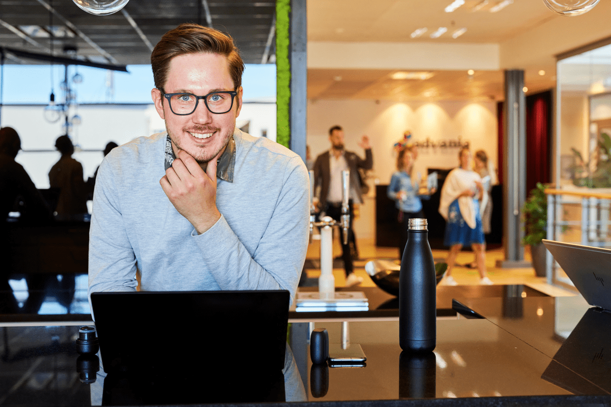 Man i blå tröja lutar sig leende över ett bord, människor rör sig i bakgrunden