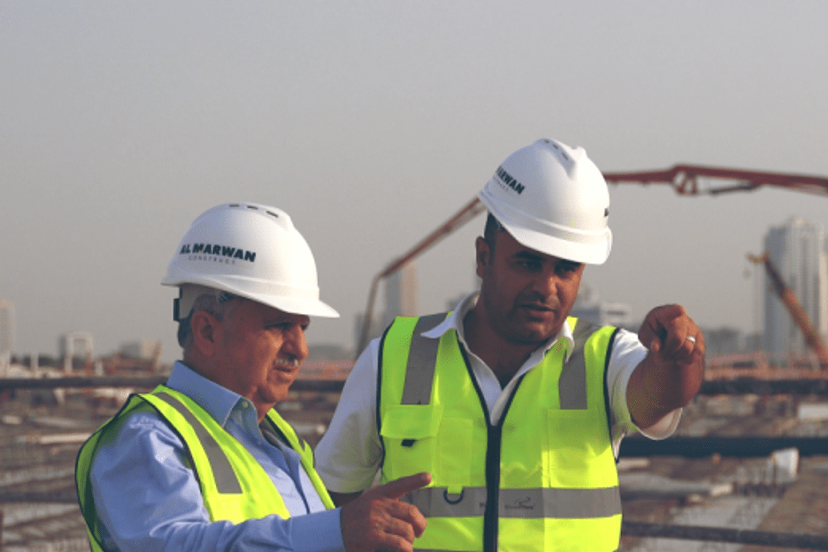 A representative from Al Marwan Group, wearing a white Kobelco-branded polo shirt, speaks with a woman at a glass table during a professional exhibition. A Gulf News newspaper and project portfolio are visible on the table.