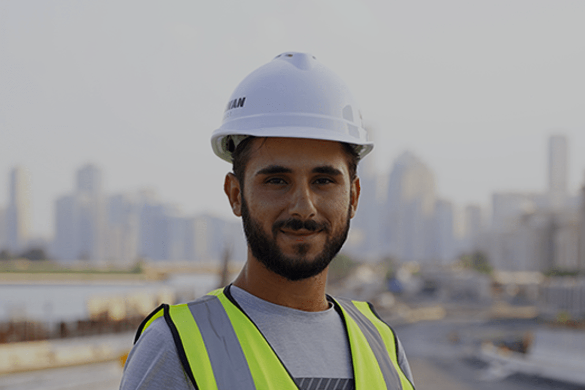 A smiling engineer from Al Marwan Construct wearing a white hard hat and yellow safety vest, holding construction plans at an outdoor site with a blurred city skyline in the background.