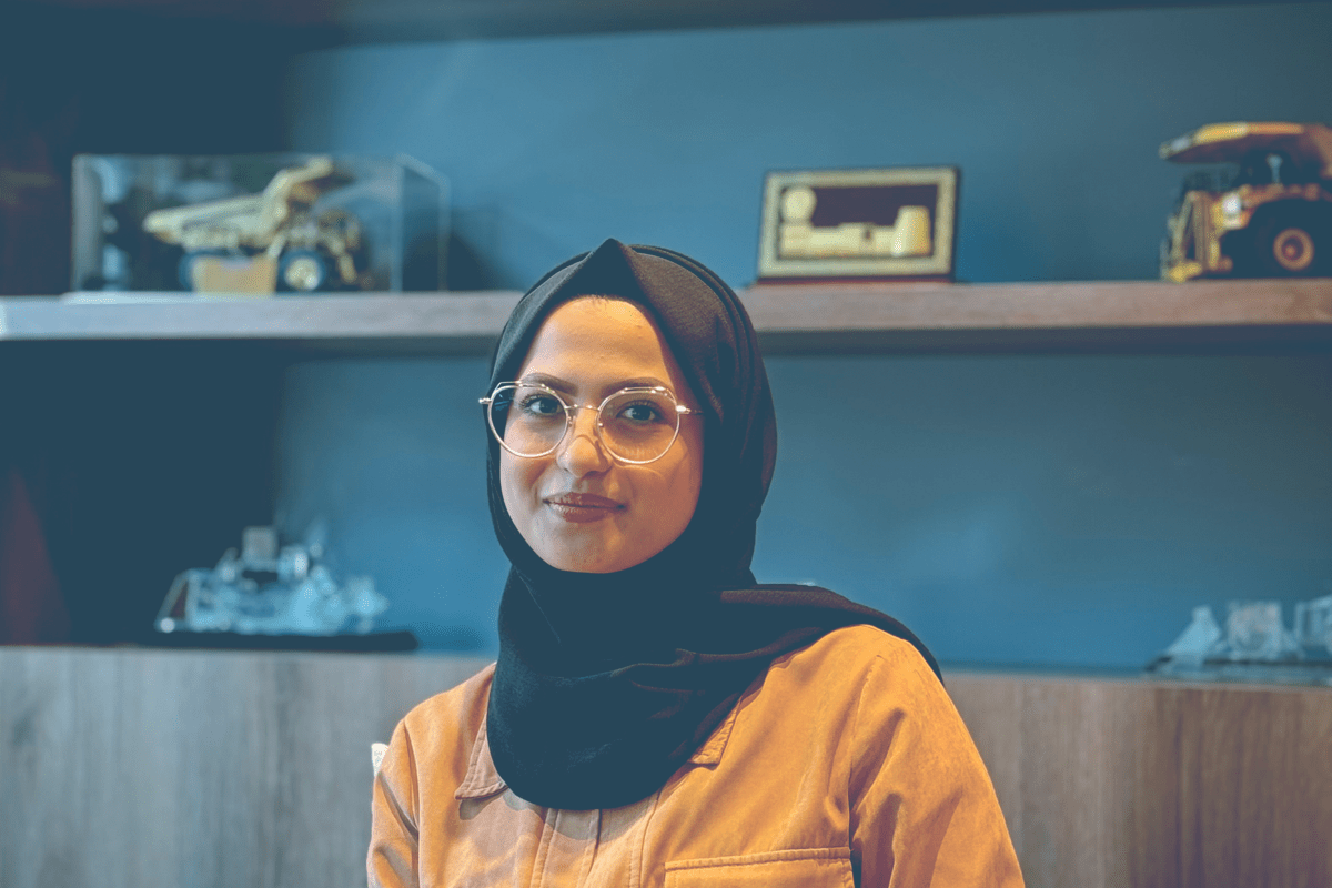 A professional woman wearing a black hijab, glasses, and a tan collared jacket sitting in a modern beige armchair. In the background, shelves display awards and scale models of heavy construction machinery.
