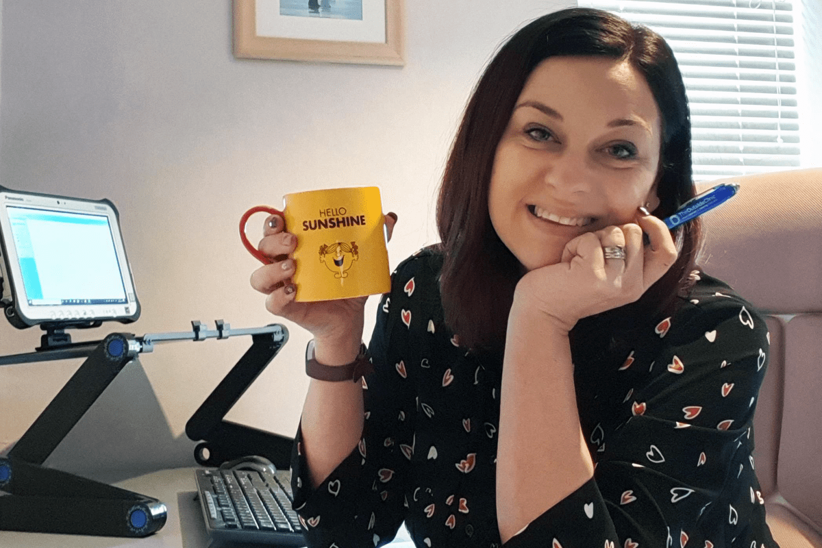 Image of woman with brown hair, sat at her desk and holding a yellow cup of tea 