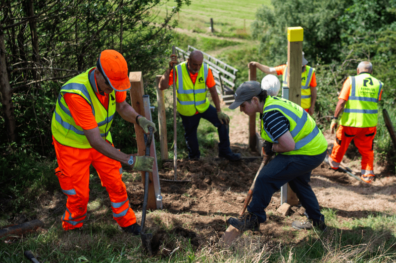 A group of volunteers wearing high-visibility clothing use spades to dig and install wooden posts on a grassy hillside. Text overlay reads "programmes and delivery".