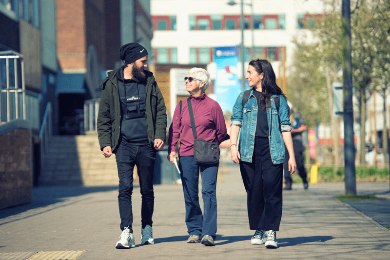 Three people walk together along a city pathway, with buildings and trees in the background. Text reads: “People & Culture”.