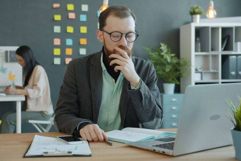 Man thinking at desk with laptop and papers.