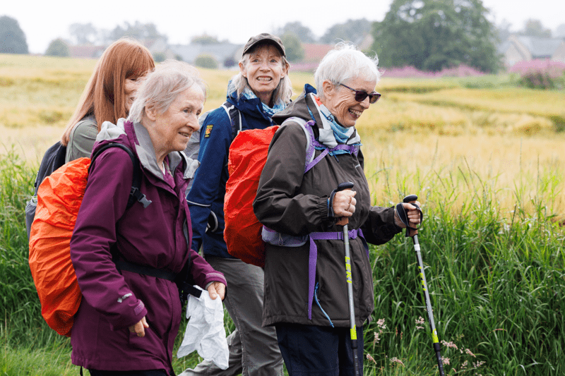 Four people wearing outdoor clothing and backpacks walk through a countryside field bordered by tall grass. One person uses walking poles, and open farmland stretches into the background. Text overlay reads “Strategy and Governance”.