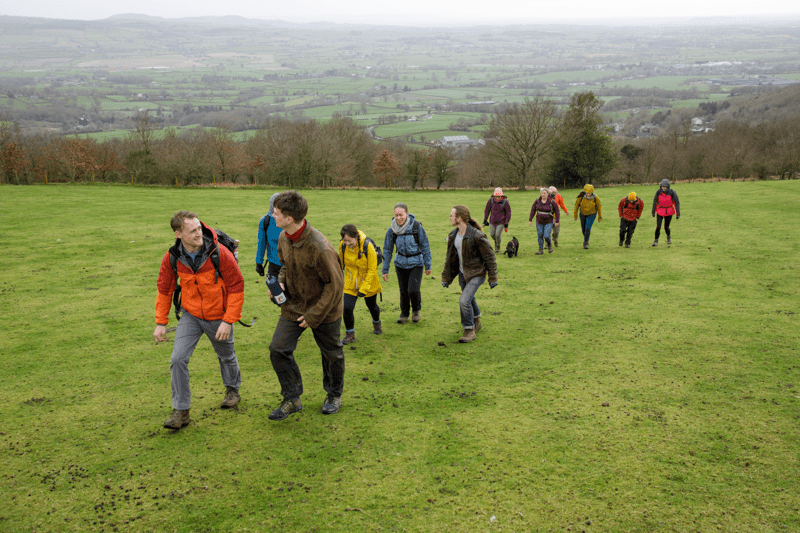 A group of walkers in outdoor clothing hike across a grassy hillside with trees and rolling countryside in the background. Overlaid text reads “Supporter Experience & Engagement” with a “Connect” button.