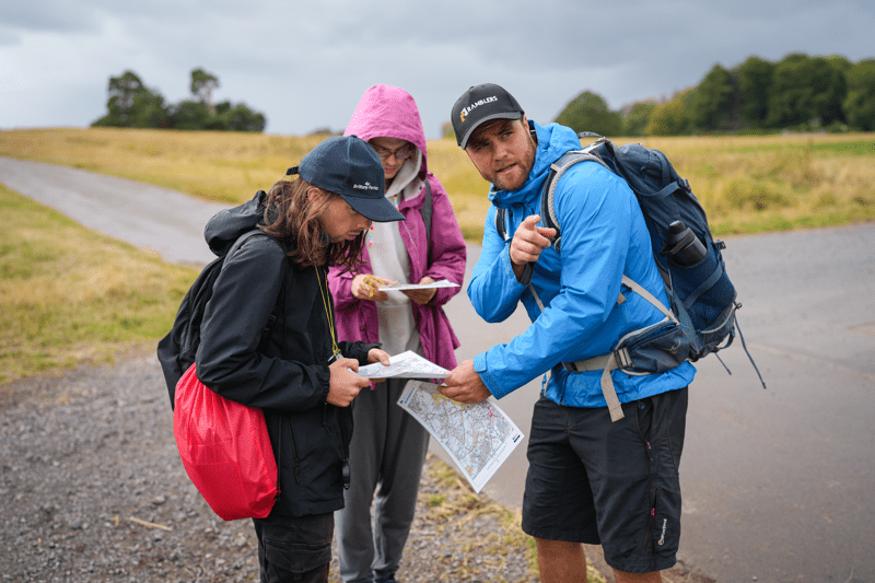 Three people in outdoor clothing stand beside a rural road, consulting maps and route notes during a walk. Text reads: “Finance”.