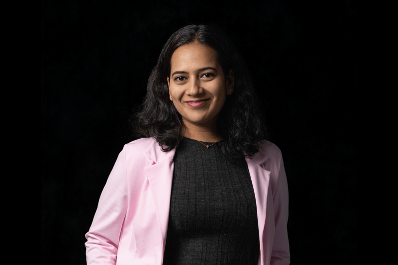 A smiling woman in a pink blazer and black outfit stands confidently against a dark background, conveying a professional and approachable tone.