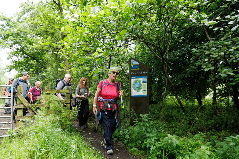 A group of walkers enter a wooded trail beside a nature reserve sign. Overlaid text reads “Fundraising.”