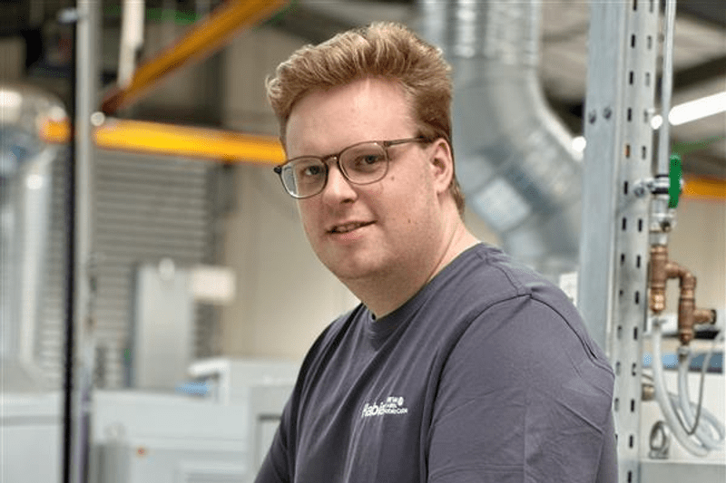 Ein Mitarbeiter in der Produktion trägt ein graues Hemd und lächelt freundlich in die Kamera.