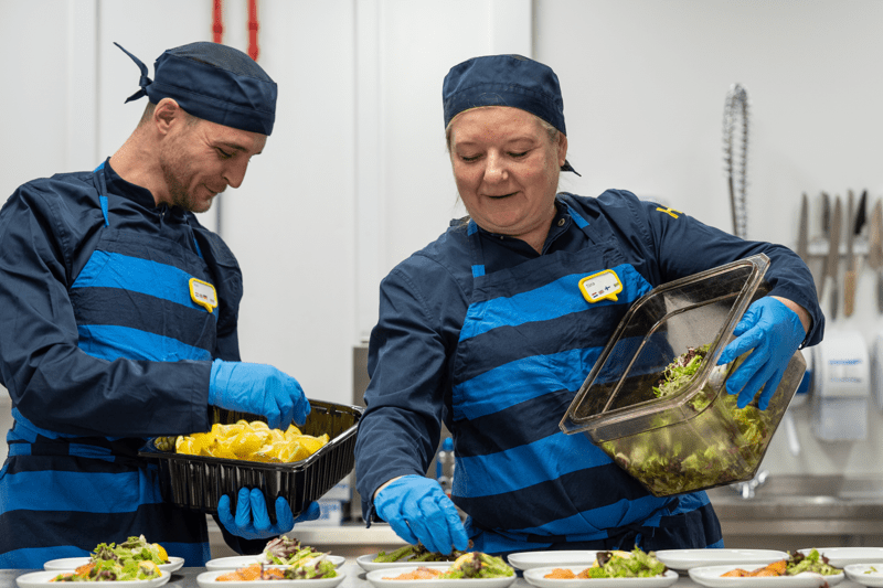 Kaks rõõmsat tumesinises vormis ja kummikinnastega IKEA köögitöötajat valmistavad salatit, asetades hoolikalt koostisaineid taldrikutele.