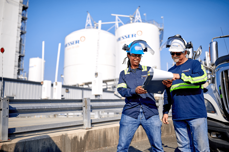 Two workers wearing helmets and protective clothing stand outdoors near industrial tanks while reviewing documents