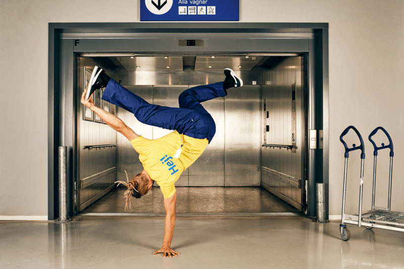 IKEA darbinieks dzeltenā uniformā balansē uz vienas rokas pie atvērta servisa lifta, labajā pusē redzami iepirkumu rati. 