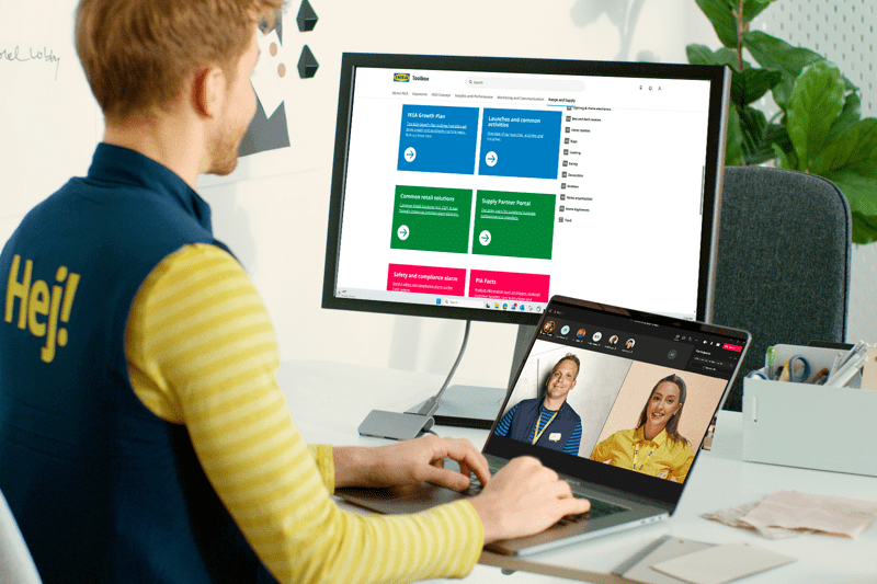 IKEA darbuotojas sėdi darbo vietoje prie nešiojamojo kompiuterio su papildomu monitoriumi ir dalyvauja vaizdo skambutyje; ant jo uniformos nugaros matosi užrašas „Hej!“