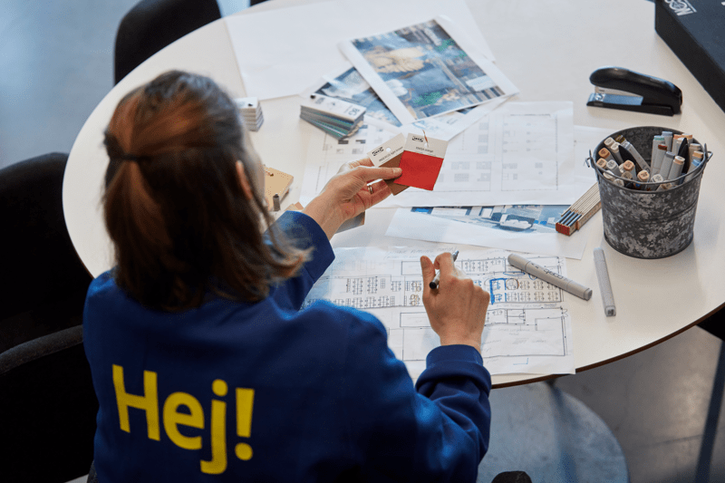 Sinises vormis IKEA töötaja, kelle jaki seljal on sõna "Hej!" istub ümmarguse laua taga, vaatab ruumiplaane ja materjalinäidiseid ning kavandab ühe toa tulevast sisustust. 