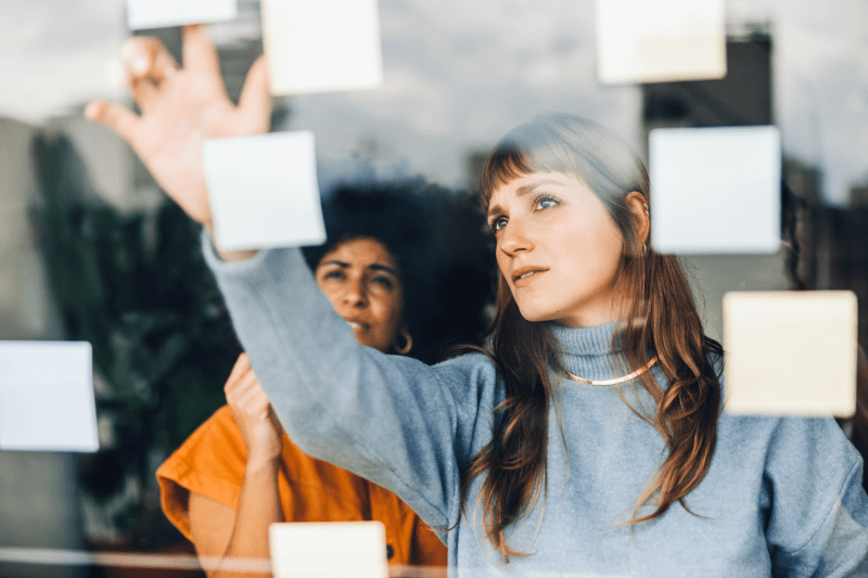 Two women standing at a glass wall, pointing at post-it notes